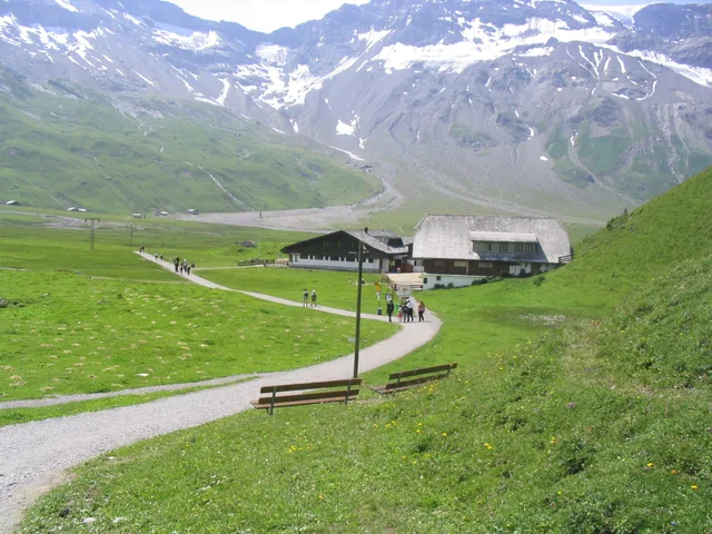 Kiesweg über die grüne Hochebene zum Berggasthaus, dahinter die schneegestreifte Felswand