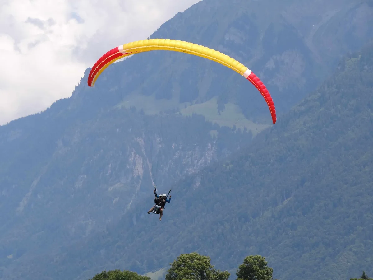 Ein rot-gelber Tandem-Gleitschirm schwebt über Interlaken vor einer bewaldeten Bergflanke