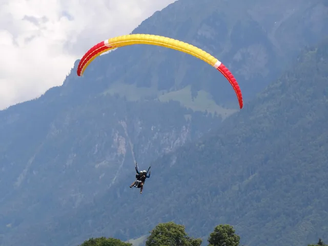 Ein rot-gelber Tandem-Gleitschirm schwebt über Interlaken vor einer bewaldeten Bergflanke