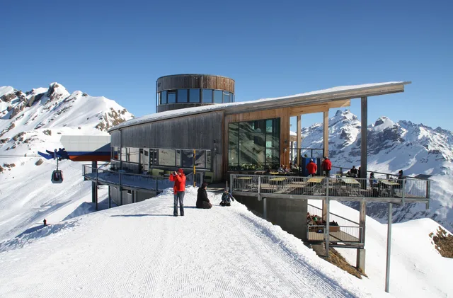 Der Alpen tower auf der verschneiten Planplatten, davor Gäste auf der Terrasse