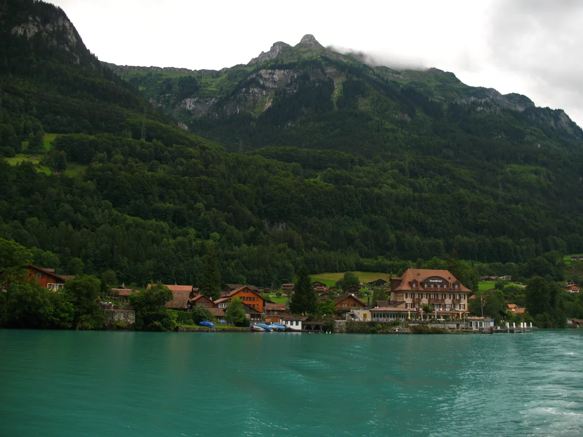 Das Dorf Iseltwald mit Holzhäusern und Hotel am Ufer des türkisfarbenen Brienzersees, dahinter ein bewaldeter Bergrücken in Wolken