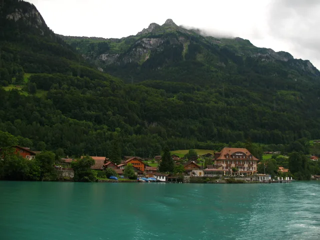 Das Dorf Iseltwald mit Holzhäusern und Hotel am Ufer des türkisfarbenen Brienzersees, dahinter ein bewaldeter Bergrücken in Wolken