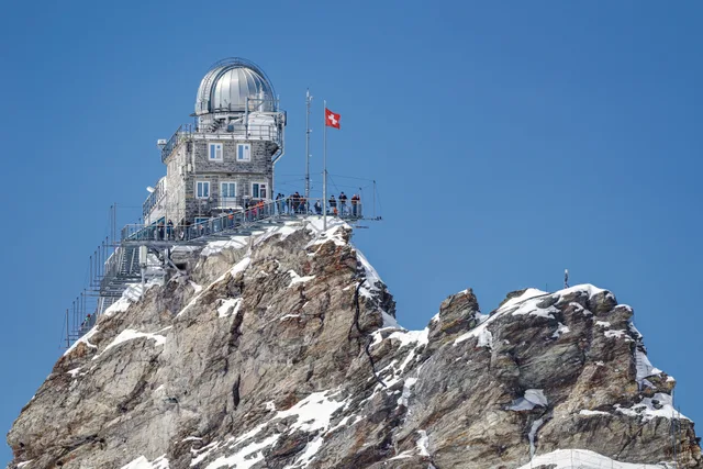 Die Sphinx-Sternwarte mit ihrer weissen Kuppel und der Schweizer Fahne auf dem Felsgrat des Jungfraujochs, darunter verschneiter Fels
