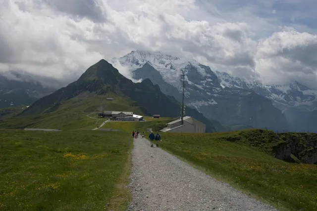 Kiesweg über den Männlichen-Grat mit Wandernden, dahinter Wolken um die Gipfel
