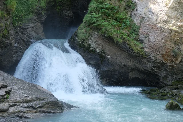 Wasserfall stürzt durch eine Felsspalte in ein türkises Becken, rechts der Steg an der Wand