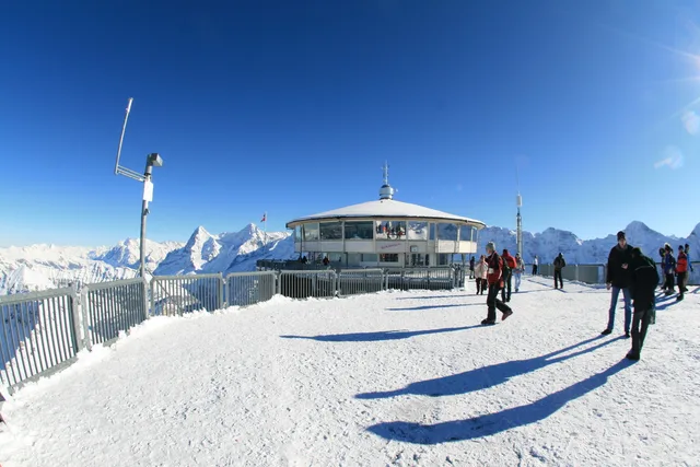 Das runde Drehrestaurant Piz Gloria auf dem verschneiten Gipfelplateau des Schilthorns, davor Besucherinnen und Besucher auf der Aussichtsterrasse