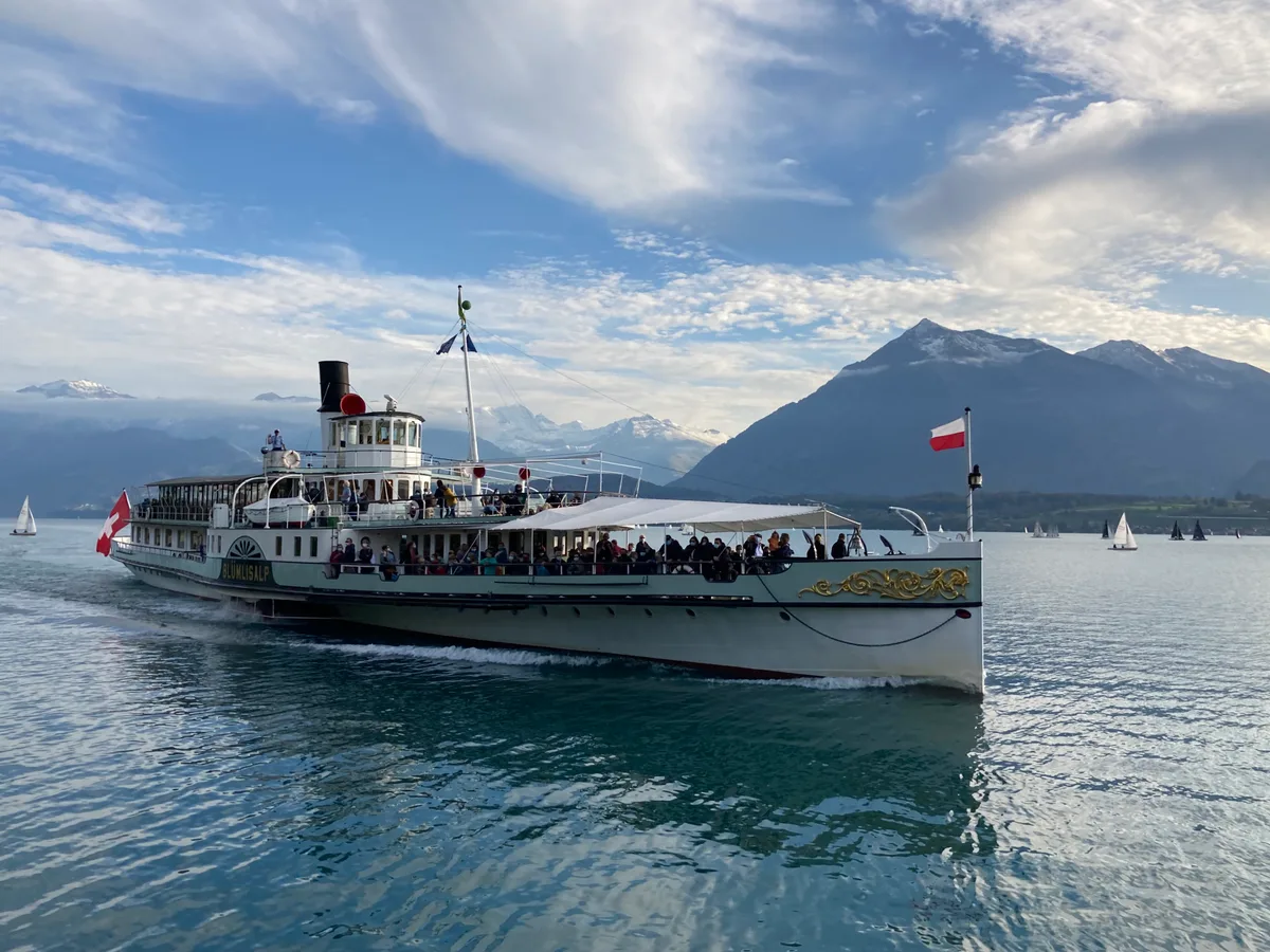 Der Schaufelraddampfer Blümlisalp in voller Fahrt auf dem Thunersee, Passagiere auf dem Oberdeck, Berge im Hintergrund