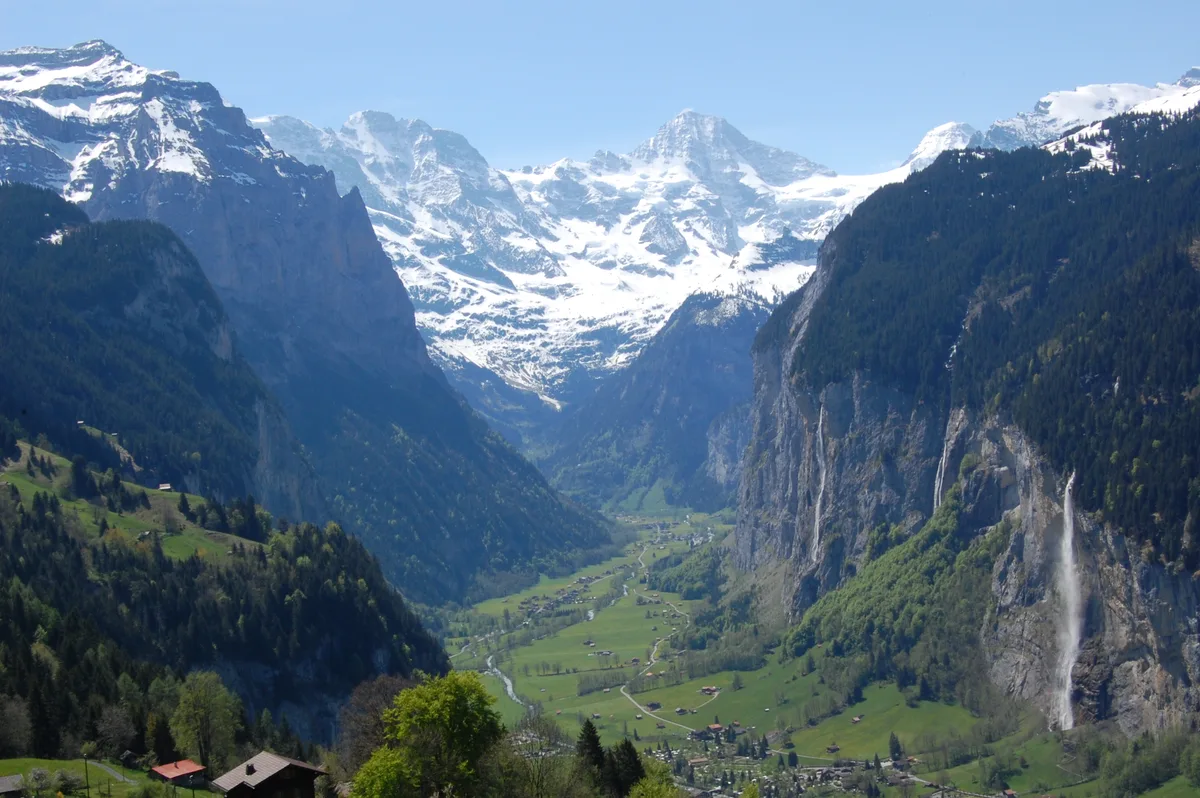 Blick von Wengen ins Lauterbrunnental, rechts ein Wasserfall über der Felswand, im Hintergrund die verschneiten Gipfel des Jungfraumassivs